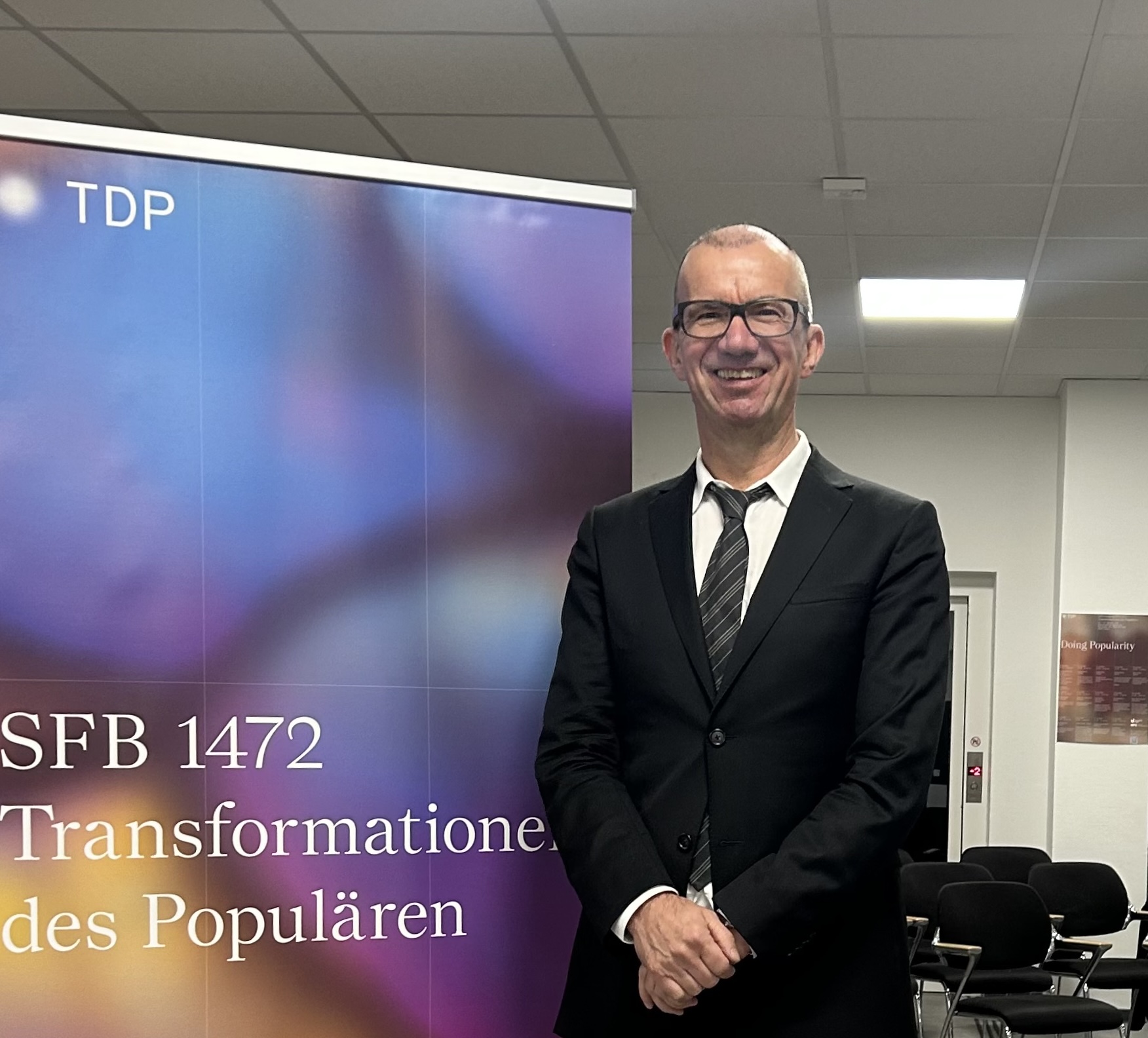 Niels Werber, schwarzer Anzug, neben einem Plakat des SFB 1472 "Transformationen des Populären"
